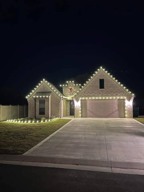 Brick home with warm white lights on gables, wreath, and lit bushes